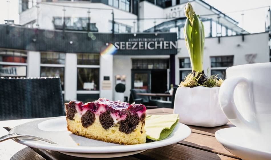 Stück Kuchen mit Kaffee auf einem Teller, serviert auf der Terrasse des Café #fraukorn im Hotel Künstlerquartier Seezeichen.