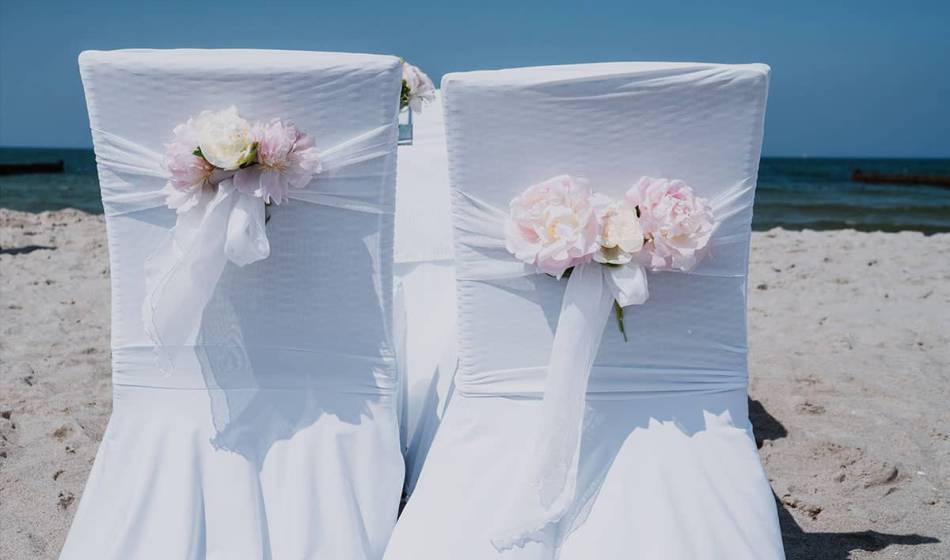 Hochzeitsstühle am Strand mit weißen Hussen und Blumendekoration, perfekt für eine Trauung an der Ostsee.