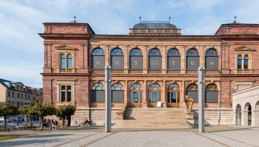 Sandsteinfassade des Museums Neues Weimar mit Rundbogenfenstern, Schriftzug „Museum“, breiter Freitreppe und goldener Skulptur, davor ein gepflasterter Platz unter blauem Himmel
