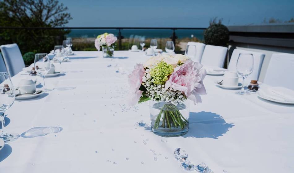 Hochzeitstafel im Freien mit weißer Tischdecke, Glasvasen mit Blumen und Ostseeblick im Hintergrund.