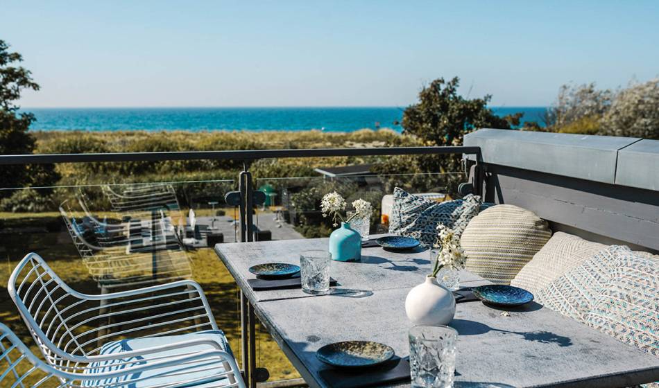 Stilvolle Terrasse im #herrhoshi in Ahrenshoop mit Tischen, Kissen und Glasgeländer, die einen Ausblick auf die Ostsee bietet.
