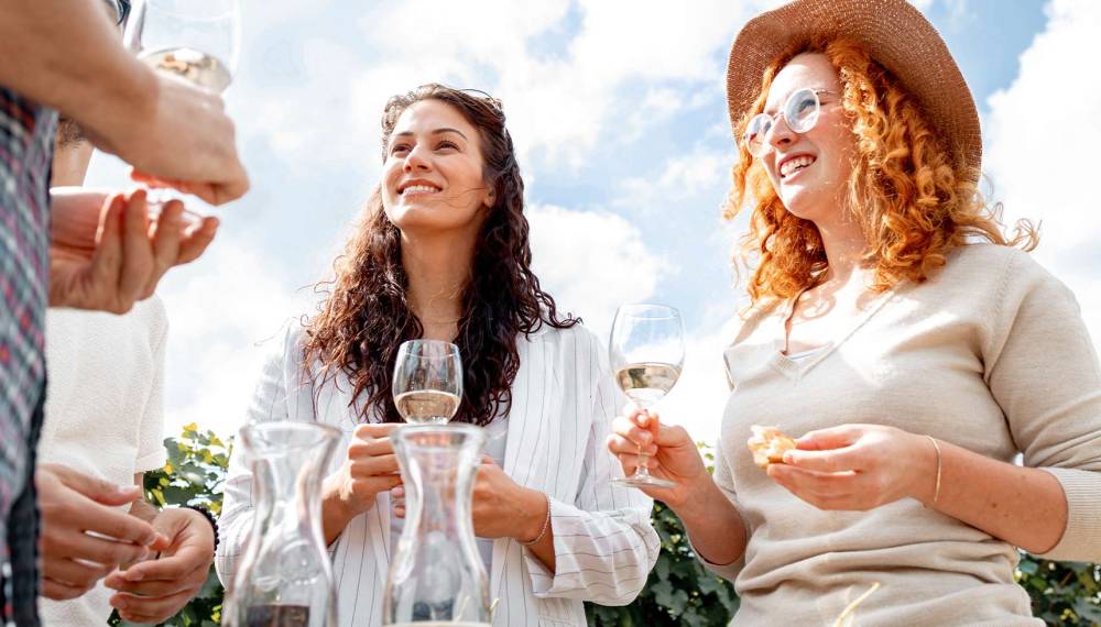 Vier Gäste genießen gemeinsam Weißwein an einem Weinstand im Sonnenschein.