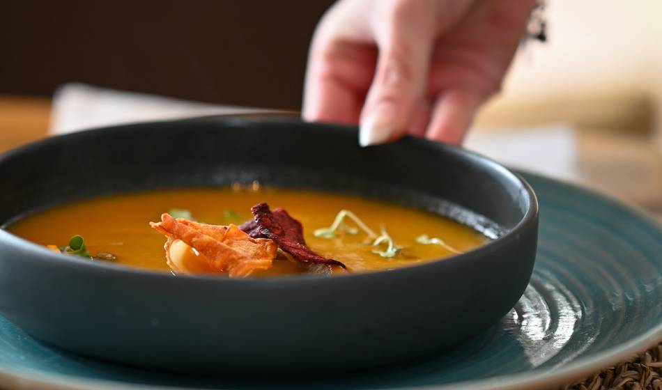 Eine schwarze Schüssel mit Suppe, dekoriert mit knusprigen Gemüsetoppings, auf einem blauen Teller im Barefoot Restaurant Mallorca.