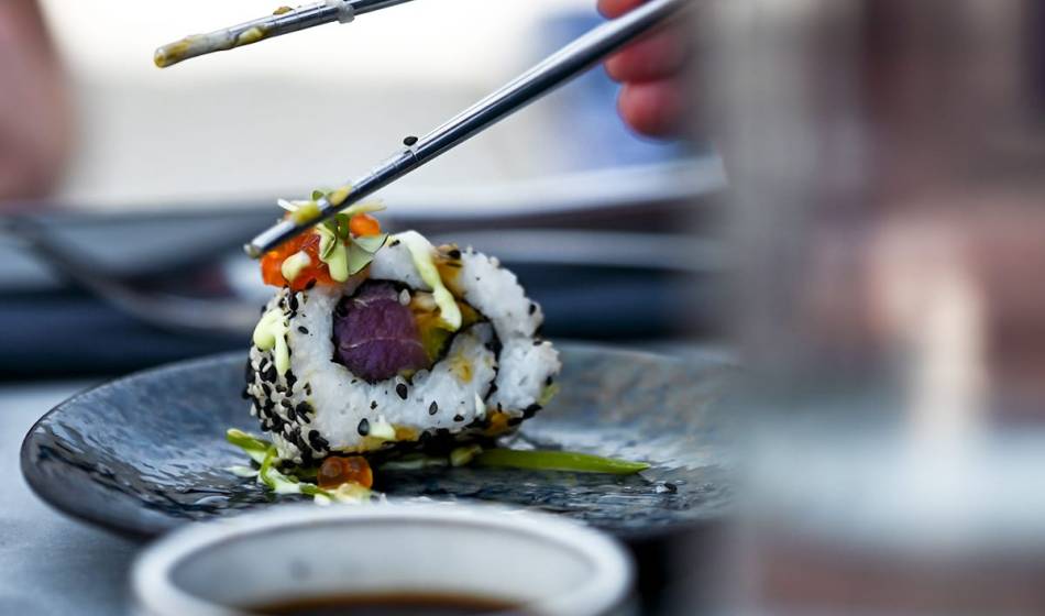 Nahaufnahme einer California Roll mit Lachs, Avocado und Sesam, gehalten von Essstäbchen über einem Teller im #herrhoshi.