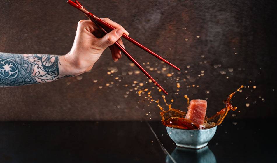 Eine tätowierte Hand mit Essstäbchen hält Sashimi über einer Schale mit Sauce, die beim Eintauchen spritzt im #herrhoshi.