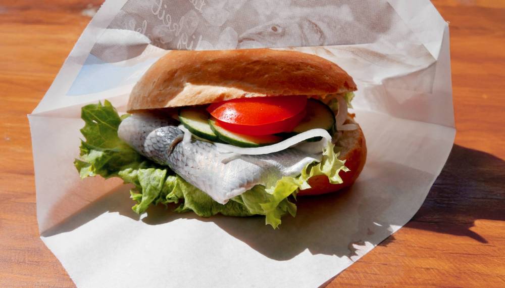 Leckeres Fischbrötchen to go in der Hand am Ostseestrand mit dem Meer und dem blauen Himmel im Hintergrund.