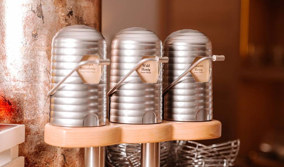 Drei metallene Honigspender mit verschiedenen Sorten, darunter Wald- und Blütenhonig, auf einem Holzständer im #fraukorn.