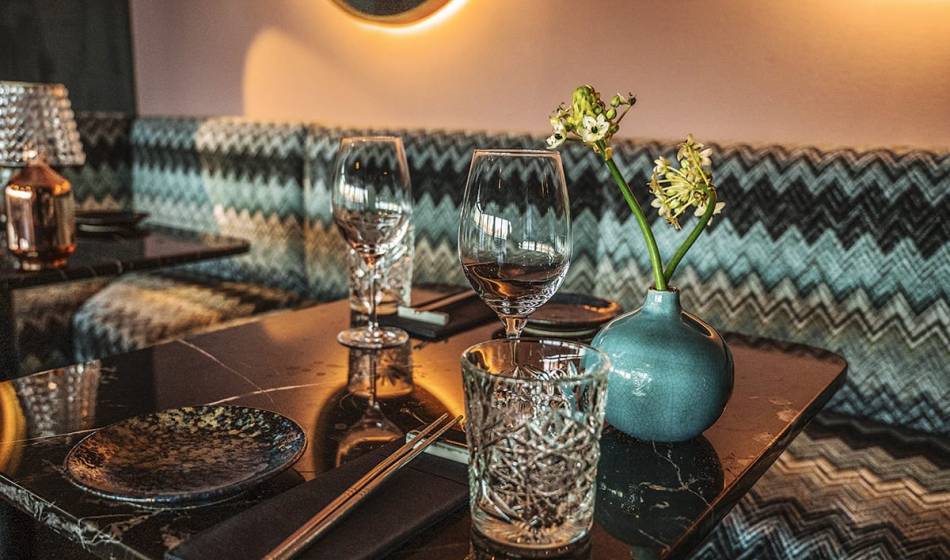 Detailaufnahme eines gedeckten Tisches im #herrhoshi mit Kristallglas, Weinglas und Vase mit Blumen, vor gemusterter Polsterbank.
