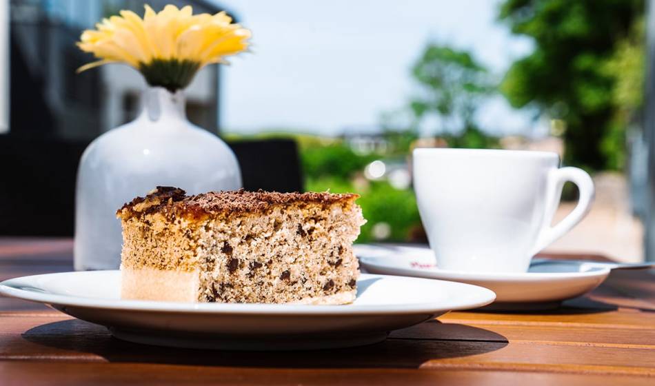 Nahaufnahme von Kuchen auf weißem Teller mit Teetasse im #fraukorn im Freien auf Holztisch mit einer Blume im Hintergrund.