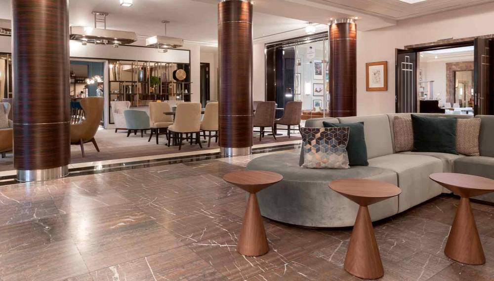 Lobby im Hotel Elephant Weimar mit Marmorboden, holzverkleideten Säulen, grauem Ecksofa und runden Beistelltischen aus Holz