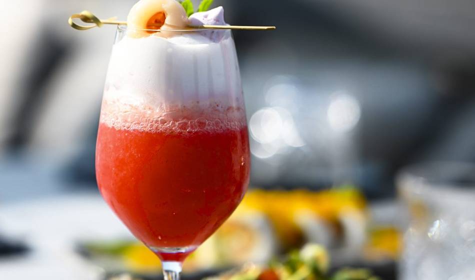 Cocktail with red juice and foam crown, garnished with lychee and mint leaf on a wooden stick at #herrhoshi in Ahrenshoop.