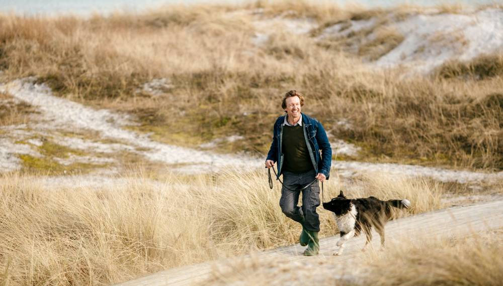 Mann spaziert mit seinem Hund zwischen Dünen und Nordsee auf Sylt.