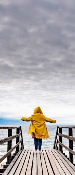 Eine Frau in einer gelben Regenjacke steht mit ausgebreiteten Armen auf einem Holzsteg am Strand von Sylt und blickt auf das Meer
