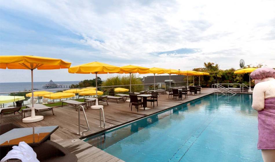 Outdoor-Pool im Kaiserhof Usedom, umgeben von Liegestühlen und Sonnenschirmen, mit der Ostsee im Hintergrund.