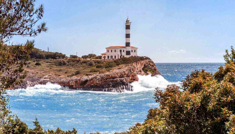 Aufnahme aus der Ferne auf den Leuchtturm von Portocolom, umgeben von einer Bucht mit klarem Meer.