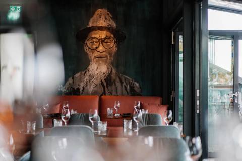 Moderner Restaurantbereich mit Holztischen, gedeckten Plätzen und Wandkunst eines Mannes mit Hut im Hintergrund.