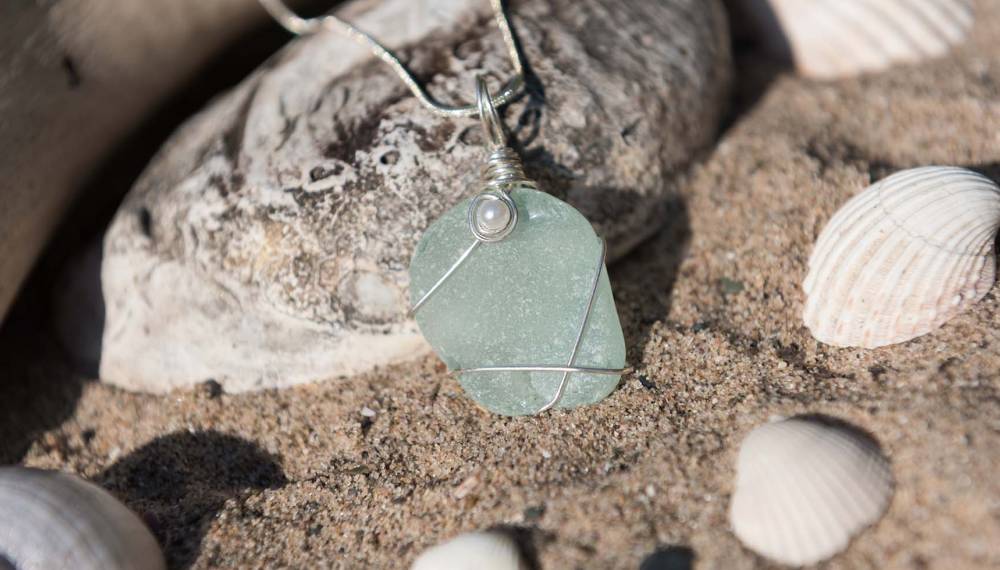 Anhänger aus grünem Seeglas mit Silberdraht auf Sand zwischen Muscheln, ein Schmuck-Mitbringsel von Rügen.