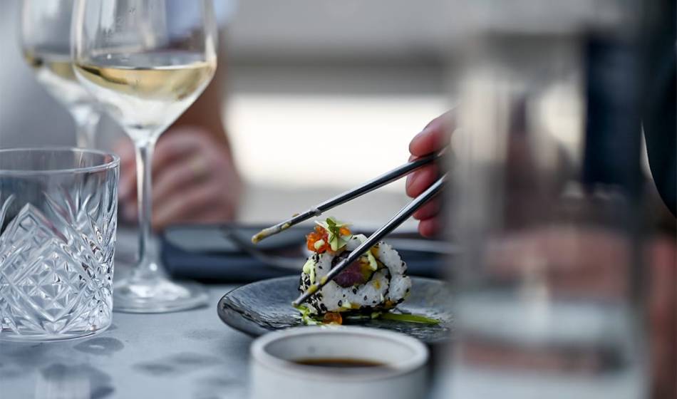 Nahaufnahme von Inside-Out-Sushi mit Sesam und Gemüsefüllung, gehalten von Essstäbchen, daneben Weingläser im #herrhoshi.