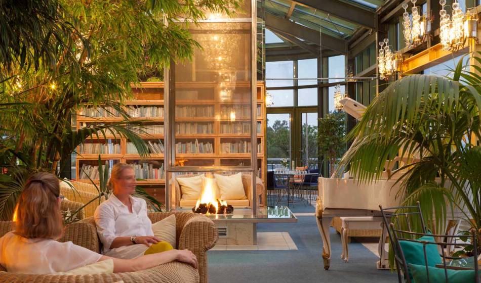 Personen sitzen im Wintergarten des Kaiserhof Usedom, umgeben von Rattanmöbeln, Kamin und Bücherregalen.