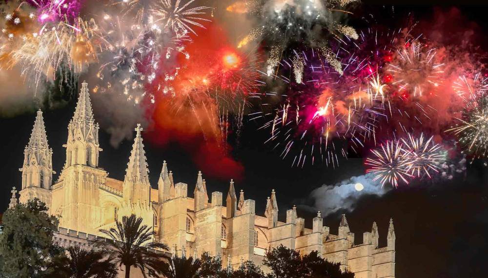 Buntes Feuerwerk am Nachthimmel über der angestrahlten gotischen Kathedrale La Seu in Palma de Mallorca mit Palmen im Vordergrund und Vollmond
