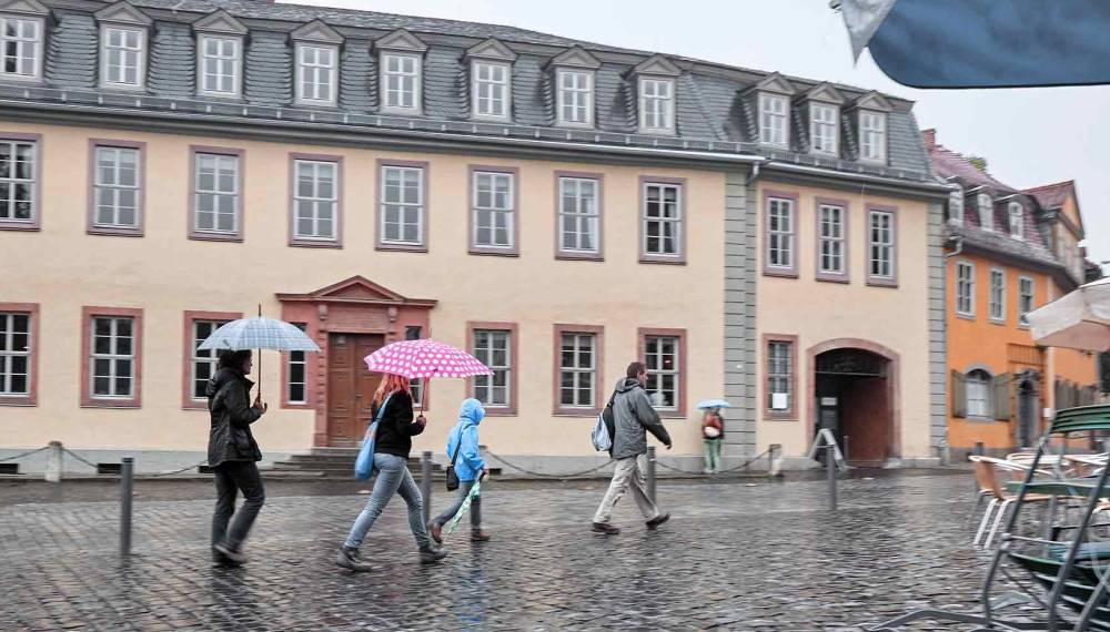 Passanten mit bunten Regenschirmen auf regennassem Kopfsteinpflaster vor der gelben Fassade von Goethes Wohnhaus am Frauenplan in Weimar, rechts gestapelte Caféstühle unter Sonnenschirmen