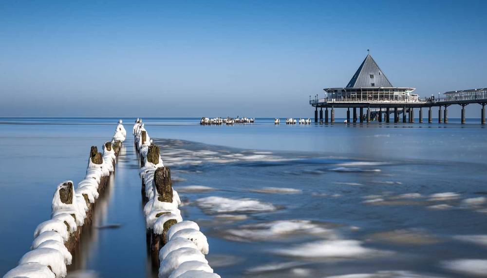 Verschneite Buhnen führen zur Seebrücke Heringsdorf über der ruhigen winterlichen Ostsee auf Usedom
