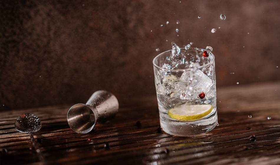 Ein Glas mit klarem Drink, Eiswürfeln, Zitronenscheibe und Wacholderbeeren, Wasser spritzt aus dem Glas im #herrhoshi.
