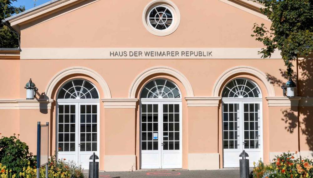 Apricotfarbene Fassade des Hauses der Weimarer Republik in Weimar mit Schriftzug, drei weißen Rundbogentüren, Rundfenster im Giebel und blühenden Beeten im Vordergrund