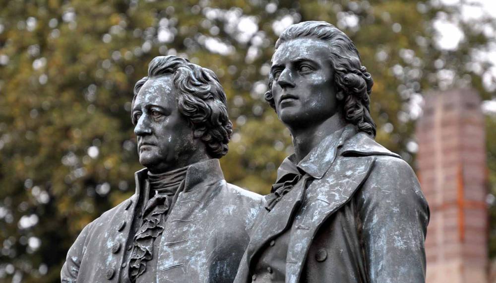 Nahaufnahme der Bronzefiguren Johann Wolfgang von Goethes und Friedrich Schillers am Goethe-Schiller-Denkmal auf dem Theaterplatz in Weimar vor herbstlich belaubten Bäumen