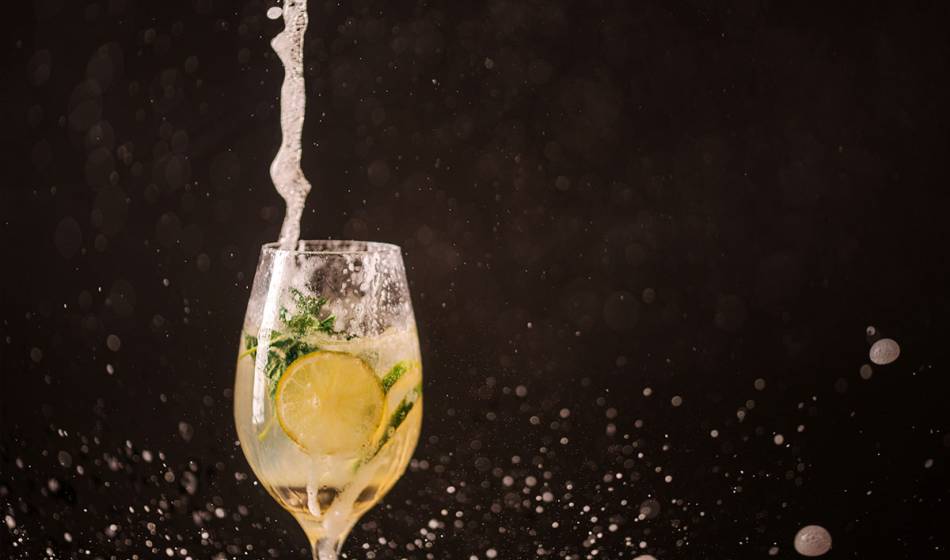 Ein Aperitifglas mit sprudelndem Getränk, Zitronenscheibe und frischer Minze, vor schwarzem Hintergrund.