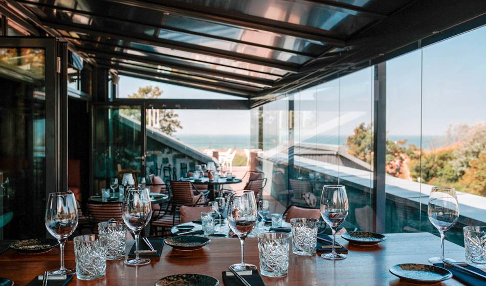 Winter garden with set tables, glasses and cutlery, surrounded by glass walls overlooking the sea in #herrhoshi.