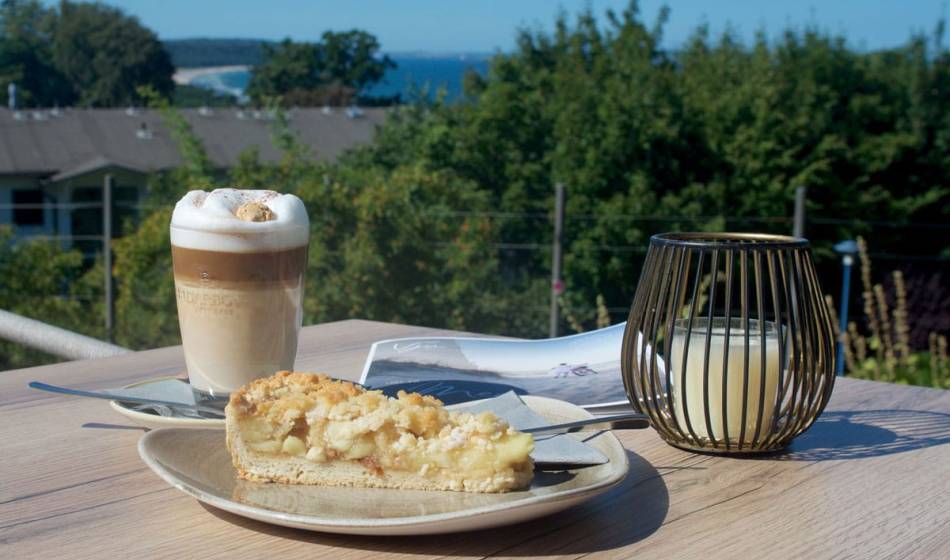 Ein Latte Macchiato und ein Stück Apfelkuchen auf einem Tisch mit Ausblick auf die Ostsee auf der See-Terasse im Vju Hotel.
