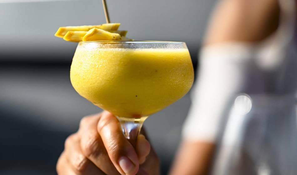 Gelber Cocktail in einem Cocktailglas, garniert mit frischen Früchten und von einer Hand gehalten im #herrhoshi in Ahrenshoop.
