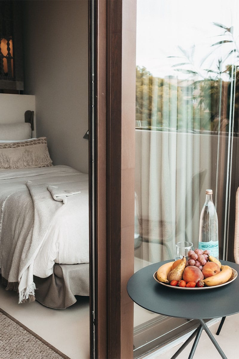 Zimmer Dormitorio im Barefoot Hotel Mallorca mit Blick auf den Balkon, Tisch mit Obstschale und Wasserflasche.