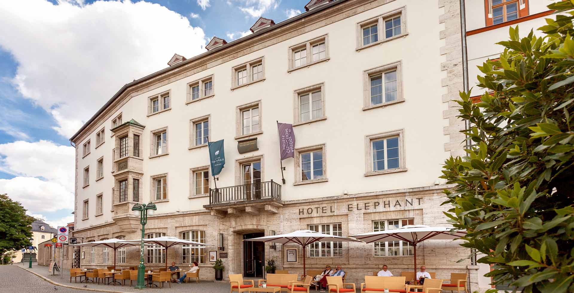 Außenansicht des historischen Hotelgebäudes des Elephant Weimar im Sommer, davor komfortable Sitzgruppen und großzügige Sonnenschirme.
