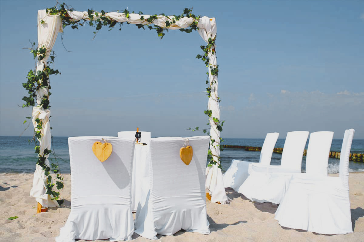 Weiß gedeckte Stühle und Traubogen am Sandstrand mit Blick aufs Meer für eine Strandhochzeit.