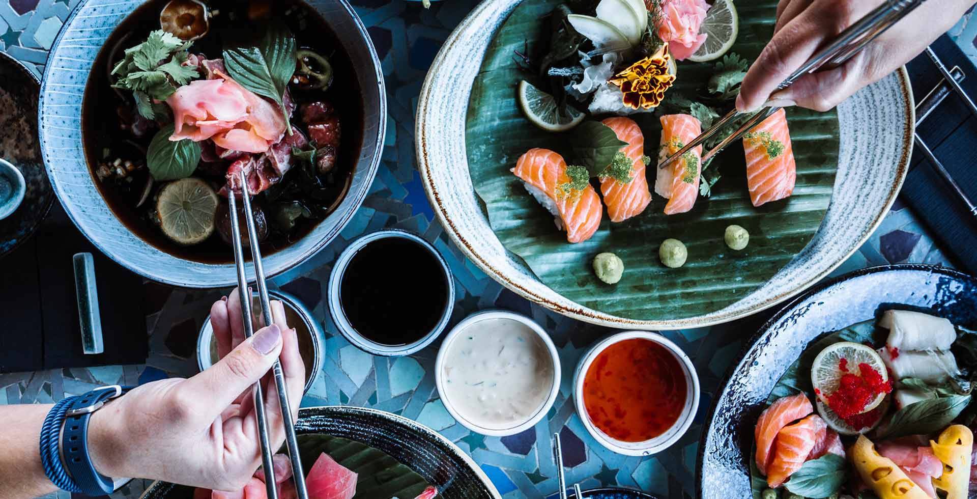 Ein üppig gedeckter Tisch mit Sushi und anderen asiatischen Spezialitäten. Zwei Personen bedienen sich davon mit Stäbchen.
