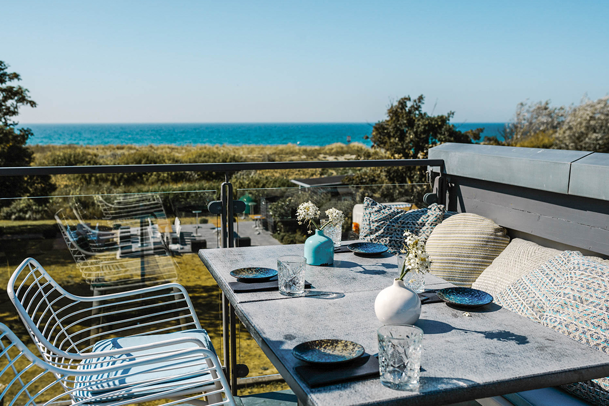 Stilvolle Terrasse im #herrhoshi in Ahrenshoop mit Tischen, Kissen und Glasgeländer, die einen Ausblick auf die Ostsee bietet.